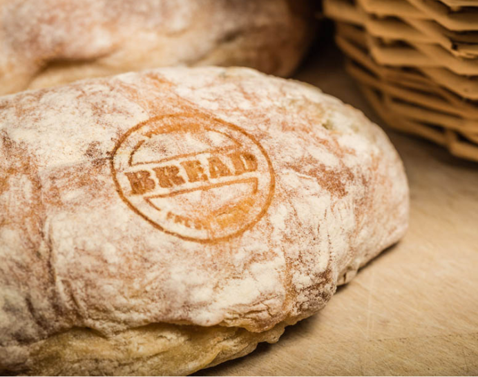Close-up of a rustic bread loaf with a laser marked circular brand logo reading "BREAD" directly on the crust