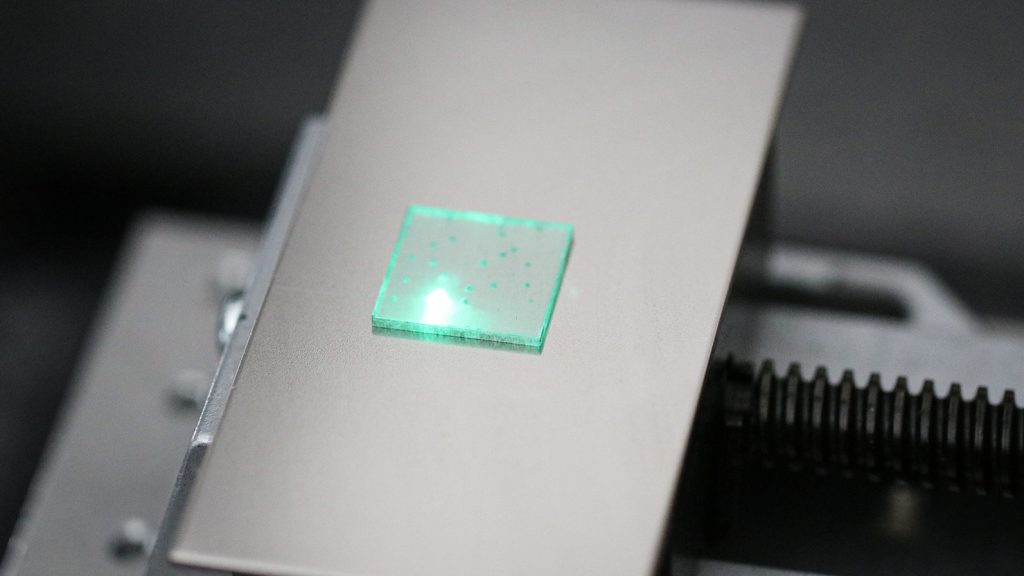 A square translucent plate with a bright green spot rests on a light-colored platform beside a black ribbed component.